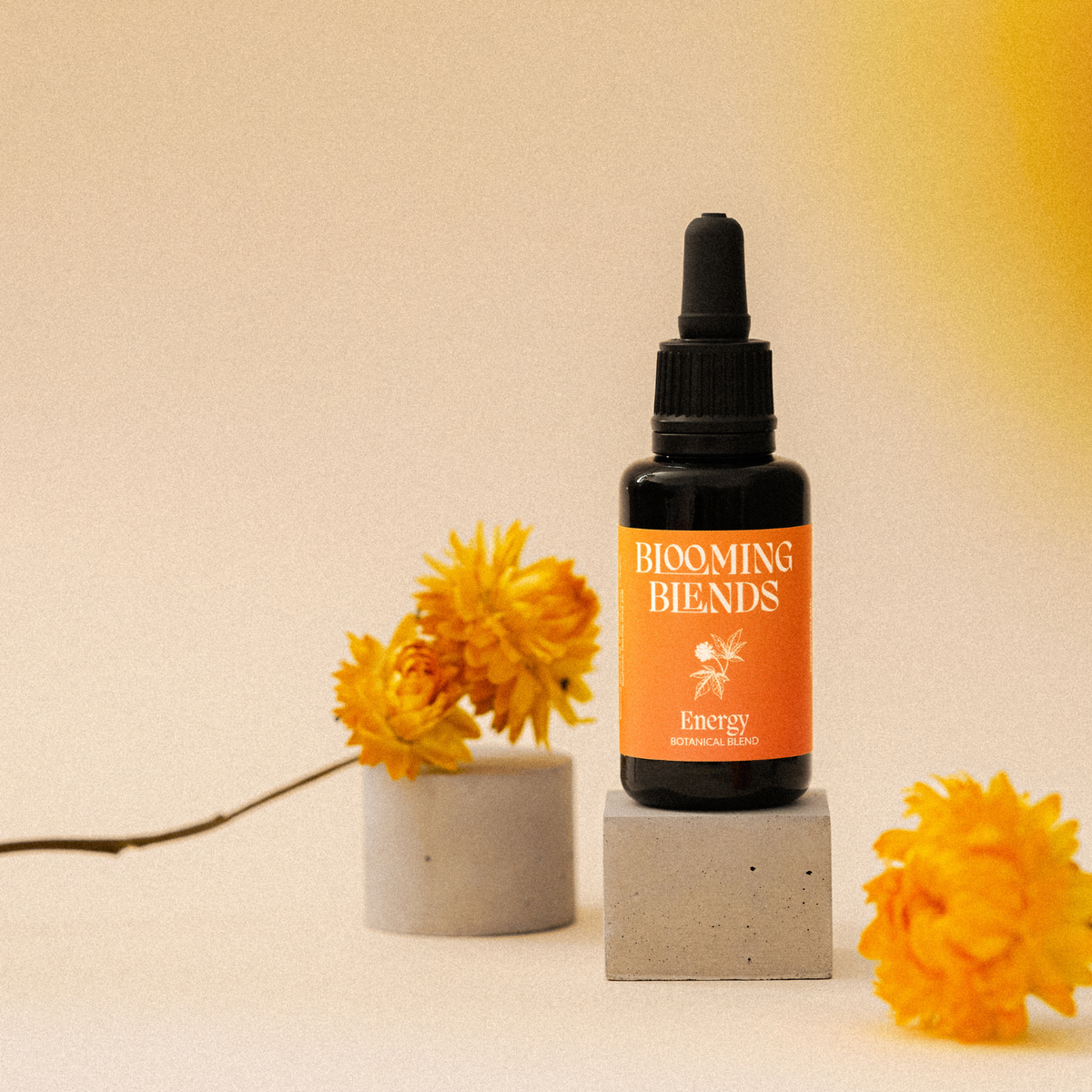 A small amber dropper bottle labeled 'BLOOMING BENDS Energy' stands on a small pedestal, styled with yellow flowers against a warm beige background.