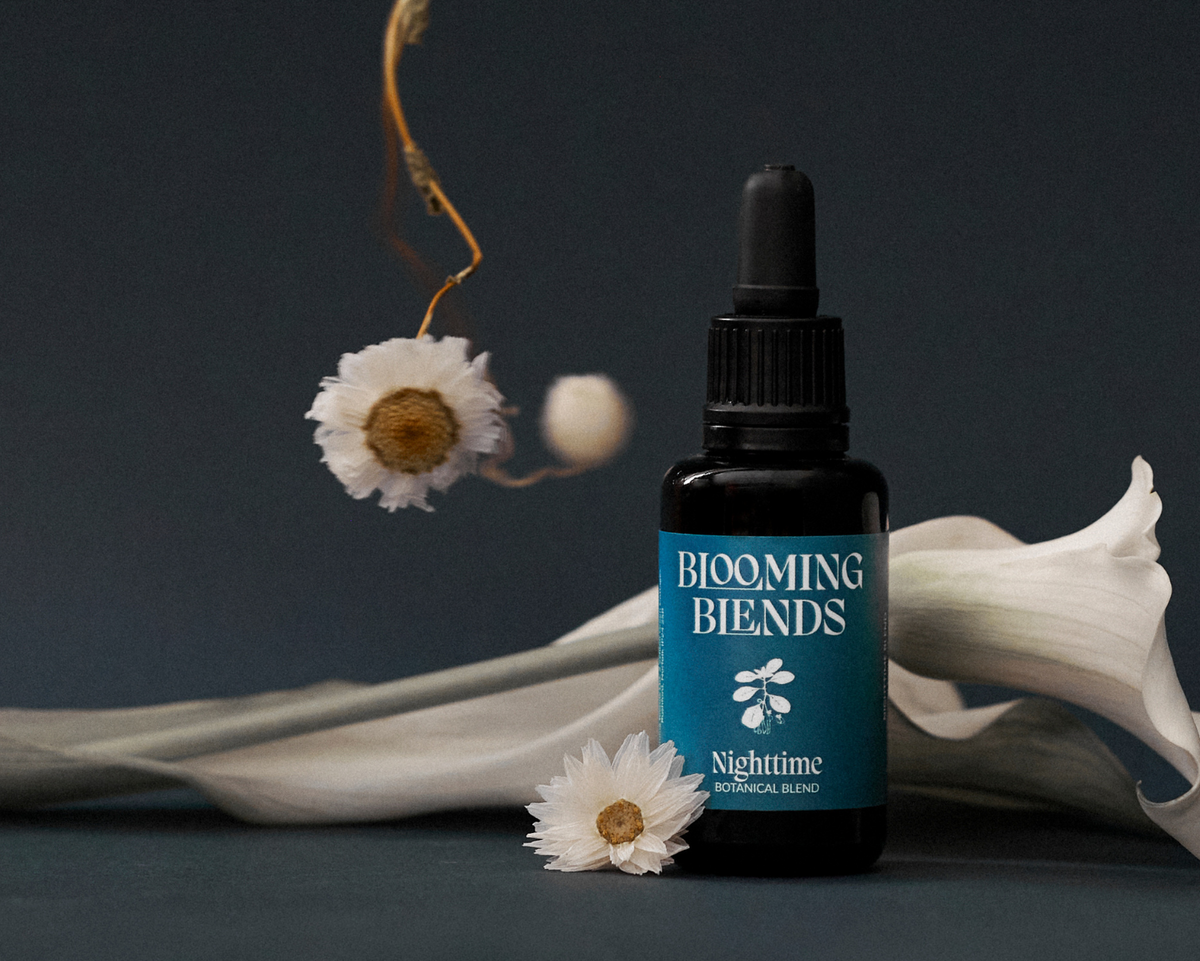 A small black dropper bottle labeled 'BLOOMING BENDS Nighttime Microbial Blend' stands on a dark surface with white flowers and a white calla lily around it against a dark charcoal background.