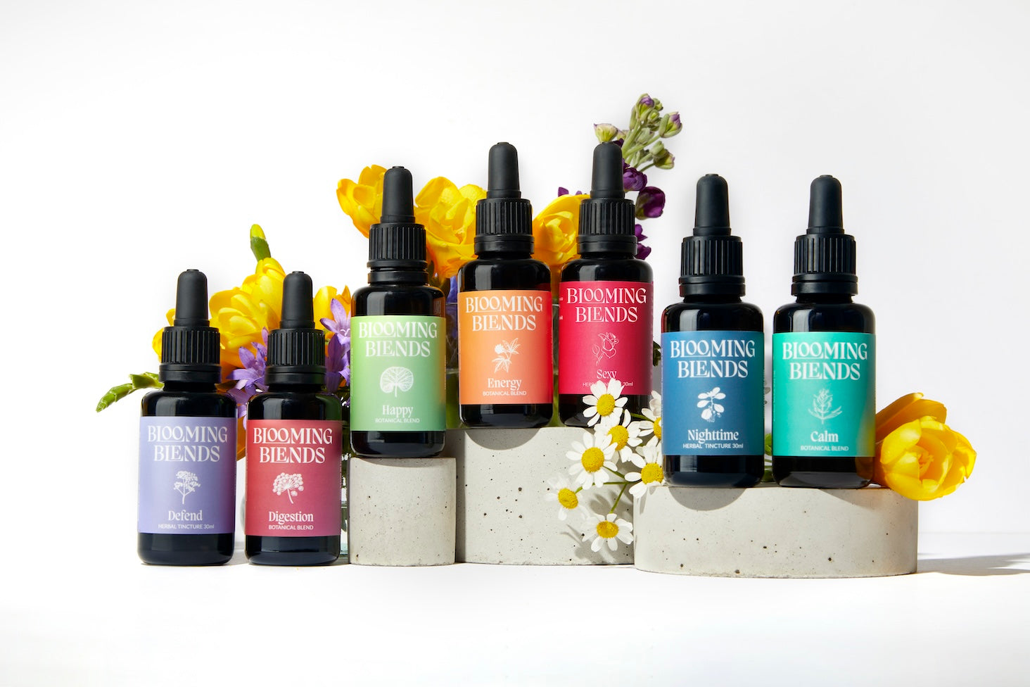 Two Blooming Blends herbal tincture bottles displayed on stone blocks with flowers.